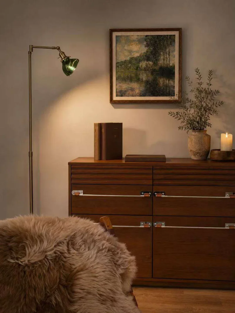 Brass floor lamp with a green glass shade beside framed landscape art and a timber sideboard.